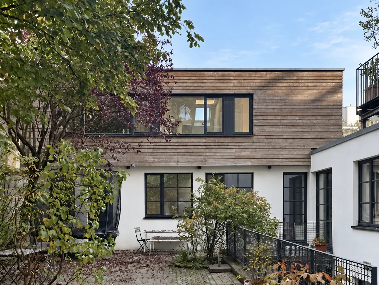 Begrünter Innenhof mit Holzaufstockung, dunklen Fenstern und Sitzbank