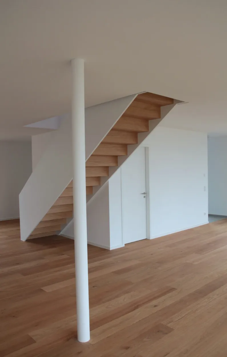 Wohnraum mit Holzboden, Holztreppe zwischen weißen Wangen und schlanker weißer Stütze, dahinter eine Tür