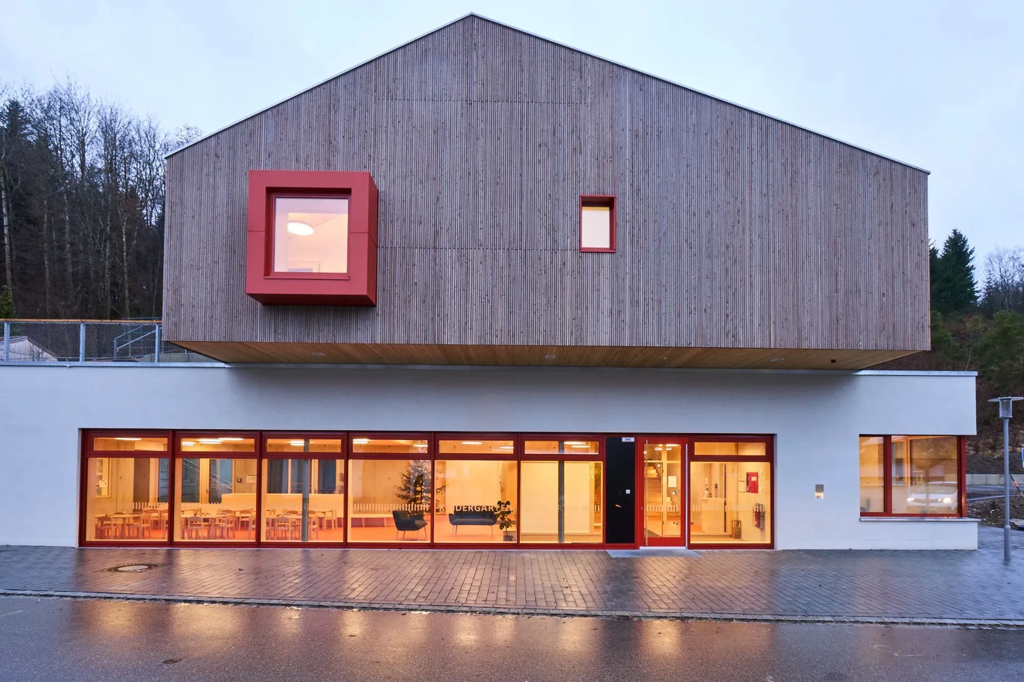 Straßenansicht des Kindergartens in der Abenddämmerung, beleuchtetes Erdgeschoss mit roten Fensterrahmen unter dem Holzgiebel