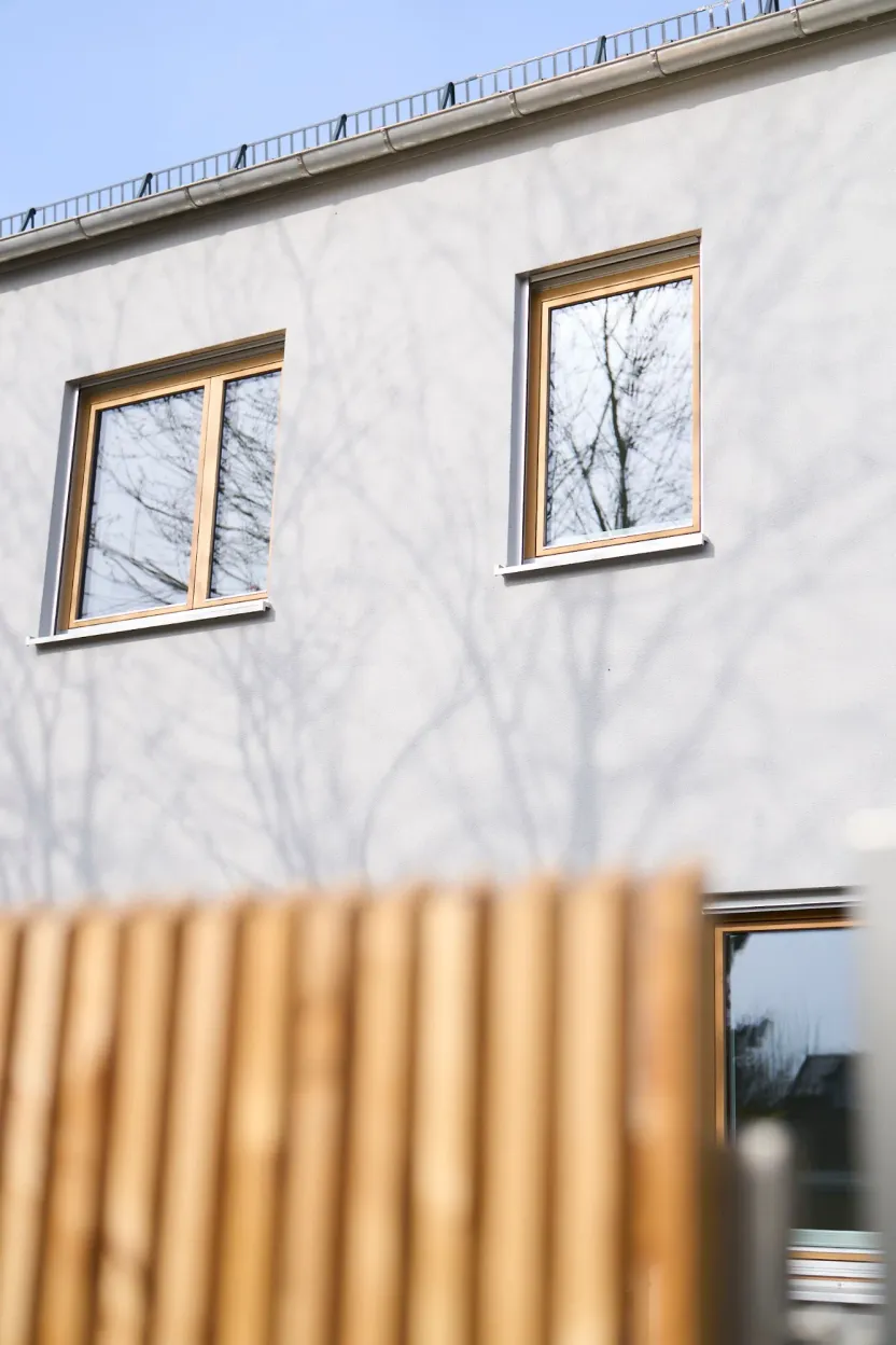 Helle Putzfassade mit zwei Holzfenstern und Baumschatten, im Vordergrund unscharf ein Holzlattenzaun