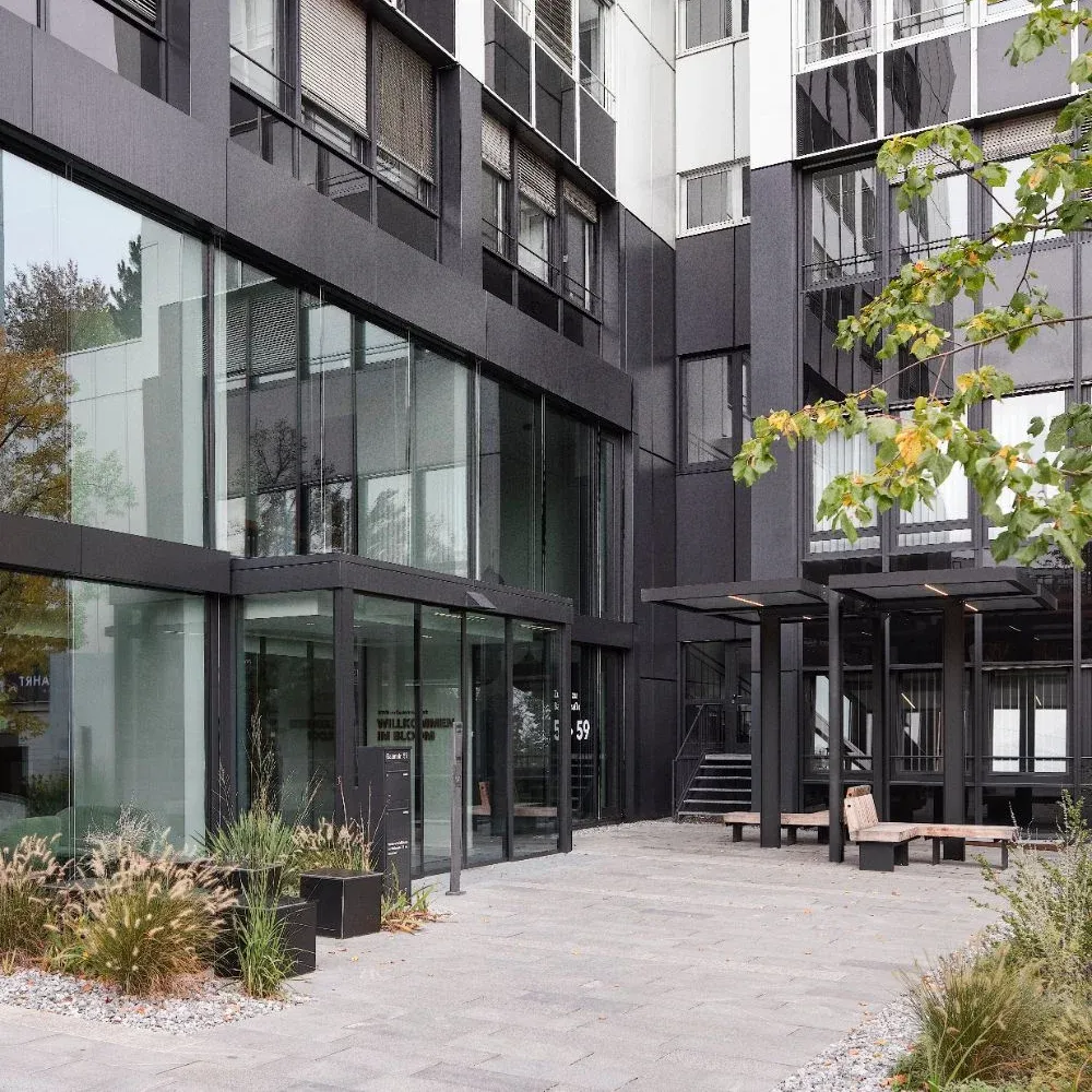 Hochhausfassade mit dunklen Paneelen und Fensterbändern über dem verglasten Eingang, davor ein Vorplatz mit Bänken