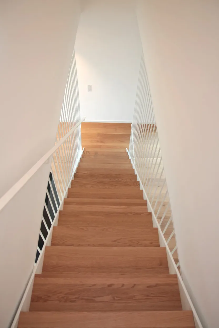 Blick von oben eine Holztreppe hinunter, beidseitig weiße Stabgeländer und Handlauf, unten Holzboden