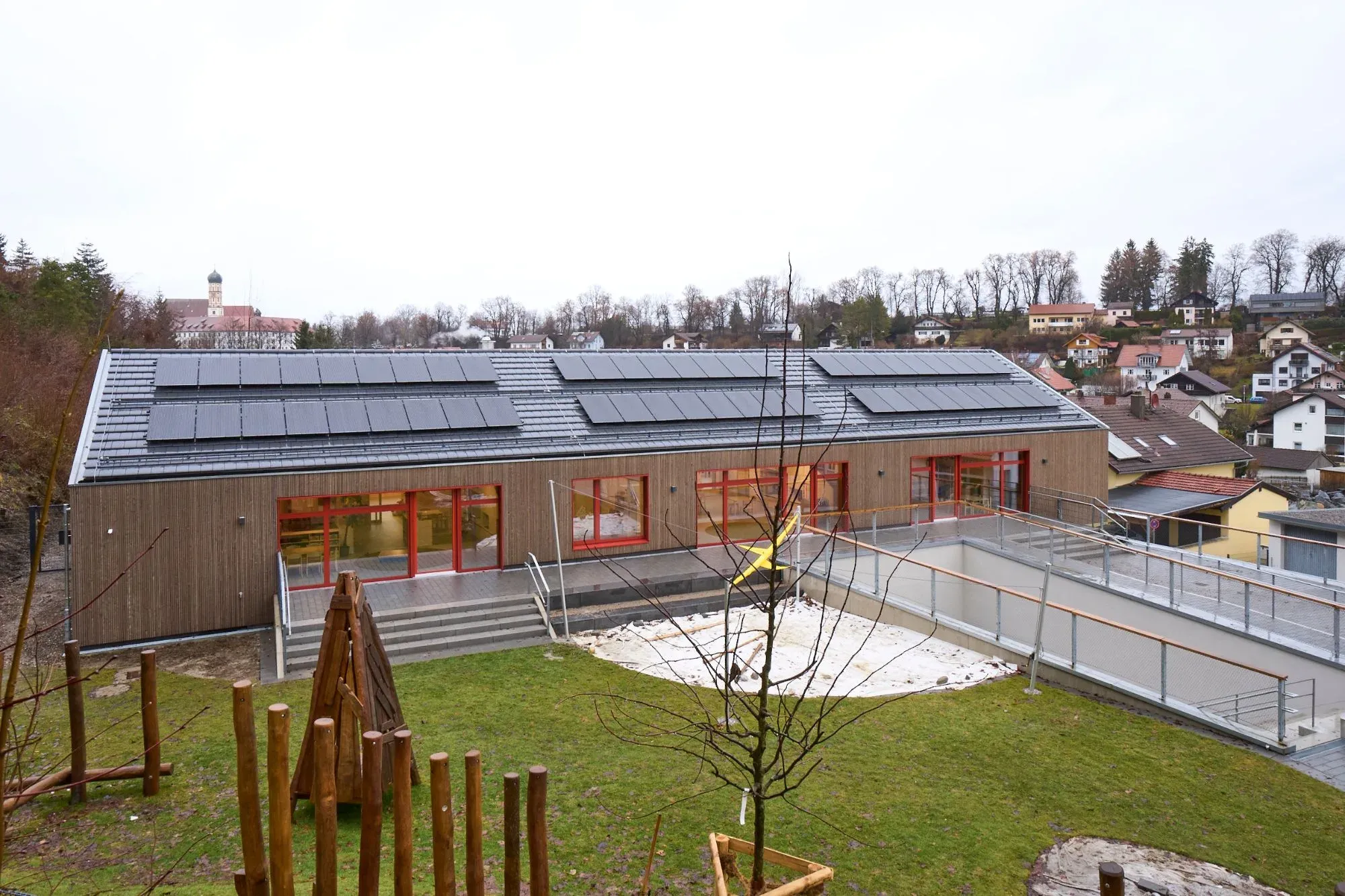 Gartenseite des Kindergartens mit Holzfassade, roten Fensterrahmen und Photovoltaikdach, davor Spielwiese mit Holzspielgeräten, im Hintergrund die Stadtpfarrkirche