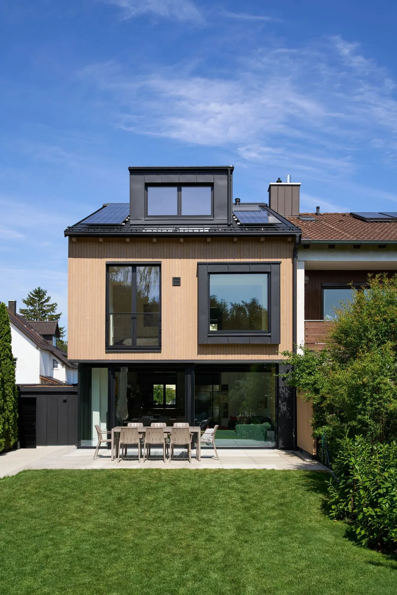 Gartenseite der Doppelhaushälfte mit heller Holzfassade, Dachgaube und Terrasse vor der Rasenfläche