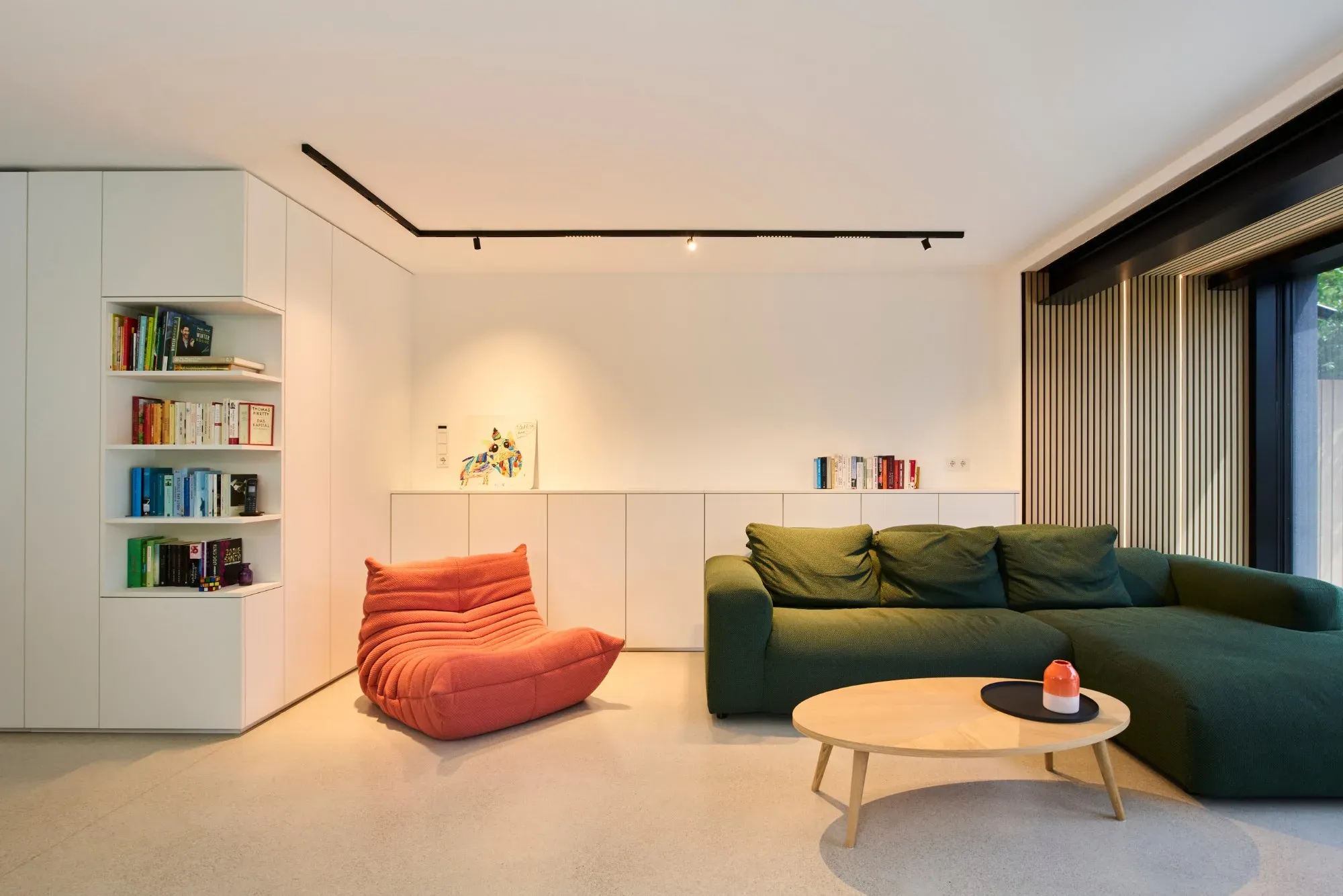 Wohnraum mit grünem Ecksofa, orangem Sessel, weißen Einbauschränken mit offenem Bücherregal und Lamellenwand am Fenster