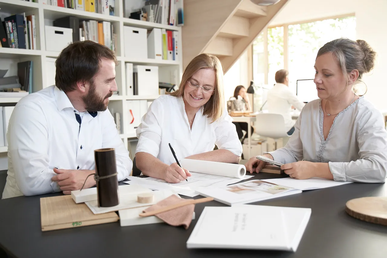 Drei Personen besprechen Zeichnungen und Materialmuster an einem Tisch im Büro von pur architekten