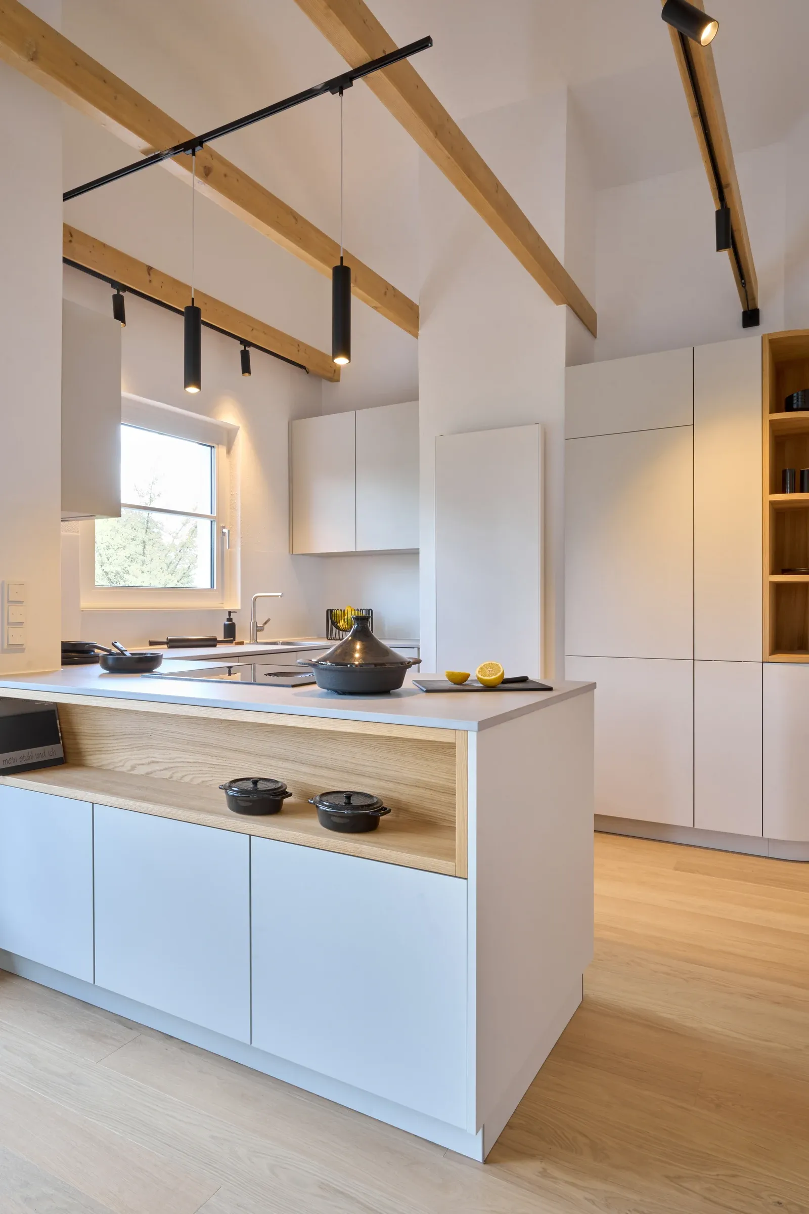 Küche mit weißen Fronten, Holzeinbauten und Eichendielen sowie eingeschalteten schwarzen Leuchten an sichtbaren Dachbalken